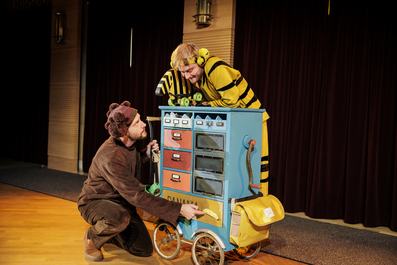 Ein als Bär verkleideter Schauspieler kniet vor einem blauen, rollbaren Schrank mit Schubladen und Fächern, während eine als Tiger verkleidete Person darauf lehnt und lächelt