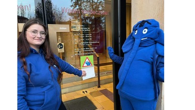 Eine junge Frau in einem blauen Hoodie steht neben einer als blauer Elefant verkleideten Person vor einer Glastür. Sie zeigt auf ein Schild mit einem Warnsymbol, das an der Tür angebracht ist.
