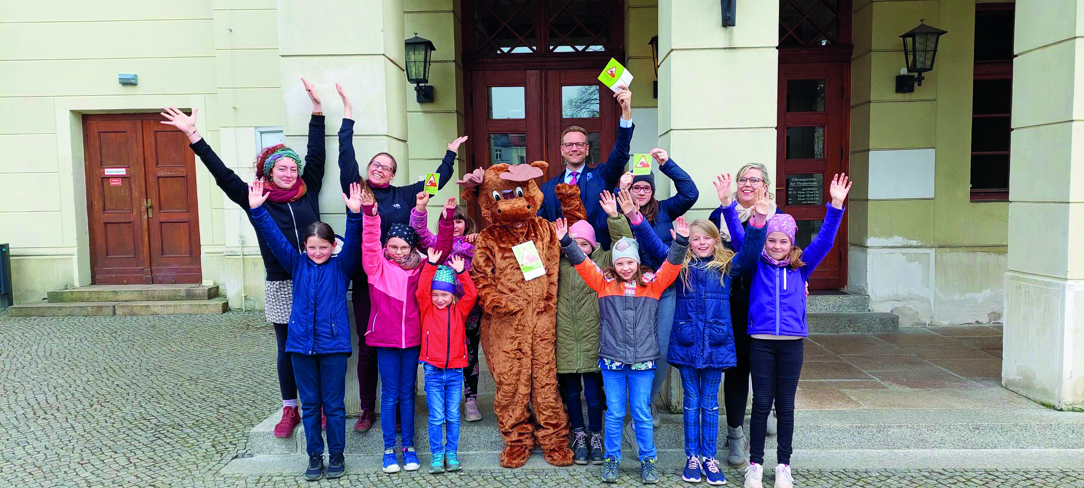 Gruppe von Kindern und Erwachsenen vor einem historischen Gebäude, gemeinsam mit einem Maskottchen in einem Elchkostüm