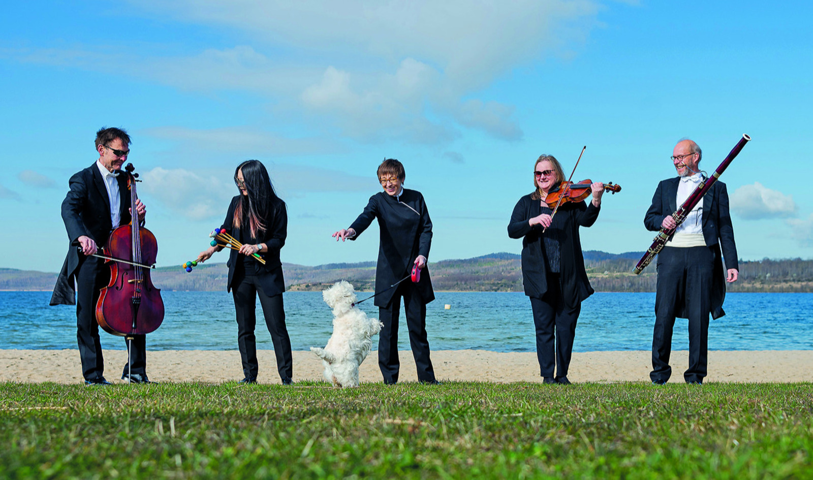 Fünf Musiker in schwarzer Konzertkleidung stehen am Ufer eines Sees mit ihren Instrumenten, während ein kleiner weißer Hund verspielt auf den Hinterbeinen steht