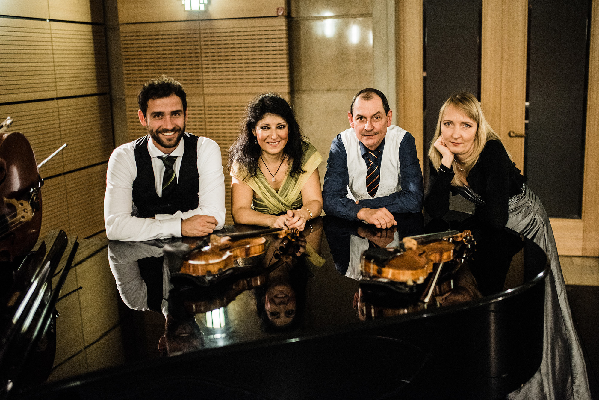 Vier Musiker und Musikerinnen sitzen lächelnd an einem Flügel, auf dem mehrere Violinen liegen. Die elegante Kleidung und das warme Licht schaffen eine stilvolle Atmosphäre