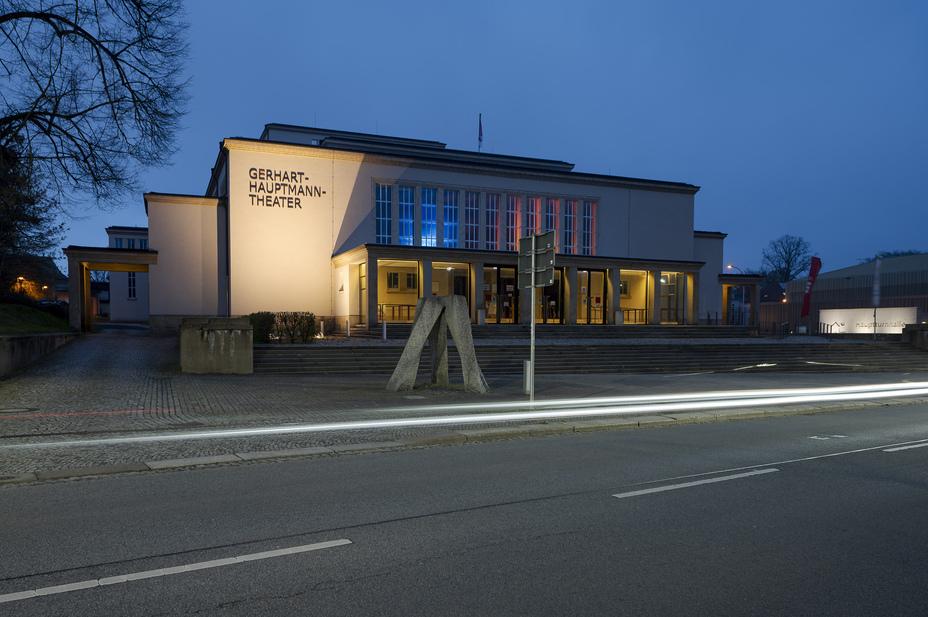 Das Gerhart-Hauptmann-Theater in Zittau bei Abenddämmerung, mit beleuchteter Fassade und Lichtspuren vorbeifahrender Autos