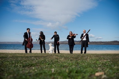Fünf Musiker in schwarzer Konzertkleidung stehen am Ufer eines Sees mit ihren Instrumenten, während ein kleiner weißer Hund verspielt auf den Hinterbeinen steht