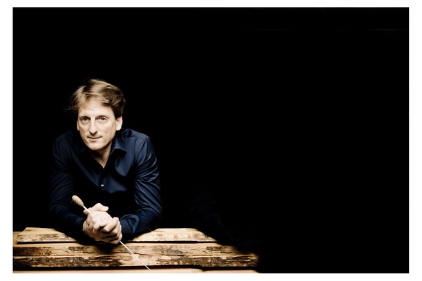 Der Dirigent Ulrich Kern in dunklem Hemd lehnt auf Holzbalken und hält einen Dirigentenstab, vor einem schwarz gehaltenen Hintergrund